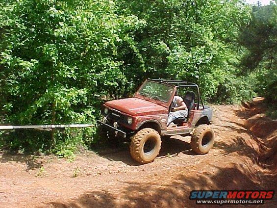 Red off-road SUV with large tires driving on a dirt trail, surrounded by trees