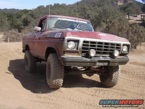 Red pickup truck with off-road tires driving on dirt, front grille and headlights visible