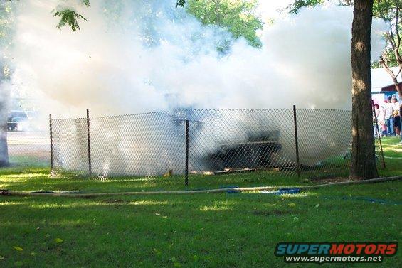 Dark vehicle obscured by smoke behind a chain-link fence