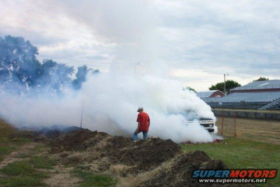 Truck blowing a lot of smoke, guy in orange shirt standing nearby. Bleachers in background