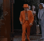 Man in an orange tuxedo with a matching hat and cane walks through a doorway, accompanied by others
