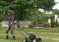 Person in a black vest and shorts mowing a lawn with a push mower