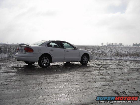White 1999 Oldsmobile Alero parked on a wet, muddy road with a field in the background