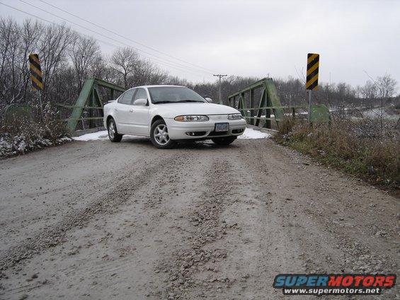 White 1999 Oldsmobile Alero on a gravel road crossing a green bridge