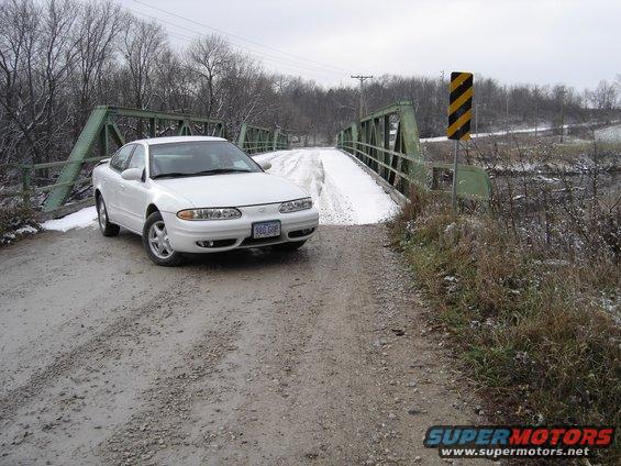 1999 Oldsmobile Alero parked on a dirt road in front of a green metal bridge, with snow on the ground
