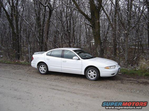 White 1999 Oldsmobile Alero parked on a dirt road with bare trees in the background