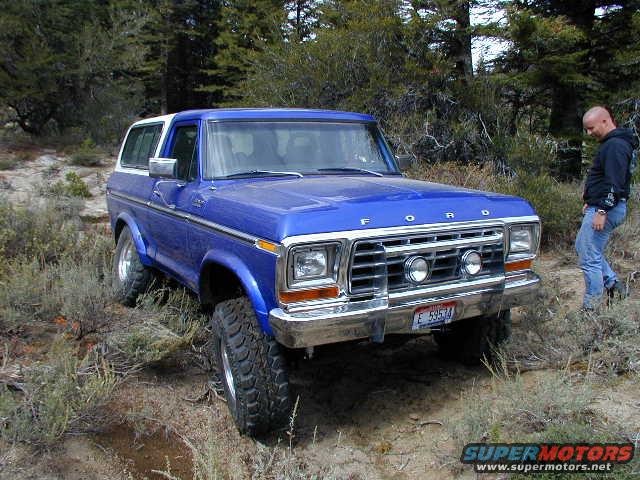 Blue 1978 Ford Bronco parked on dirt road with trees in background. Man stands to the right