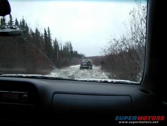 View from inside a car looking at another SUV driving on a snowy, icy road with trees on either side