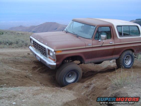 Brown 1978 Ford Bronco with a white top driving off-road, front-left wheel in a ditch