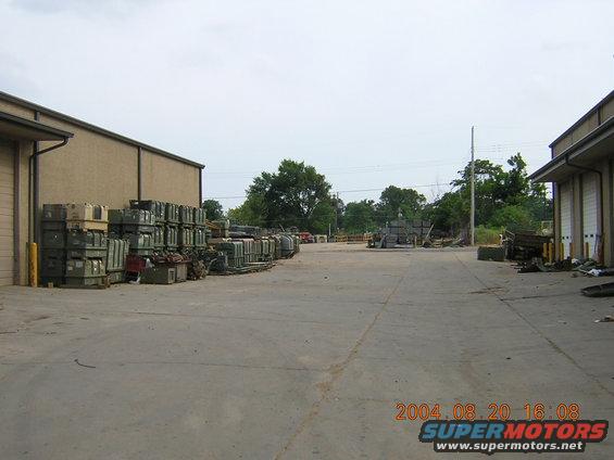 Storage yard with stacked crates next to buildings, some vehicles in distance