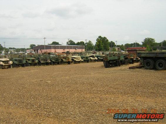 Many military trucks parked in an outdoor lot with a building in the background
