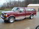 1996 Ford F-150 - A maroon 1996 Ford F-150 pickup truck is parked on a concrete surface with a wooded area and a building in the background.