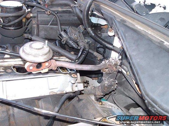 Engine bay showing a damaged exhaust pipe with signs of a leak and surrounding wiring