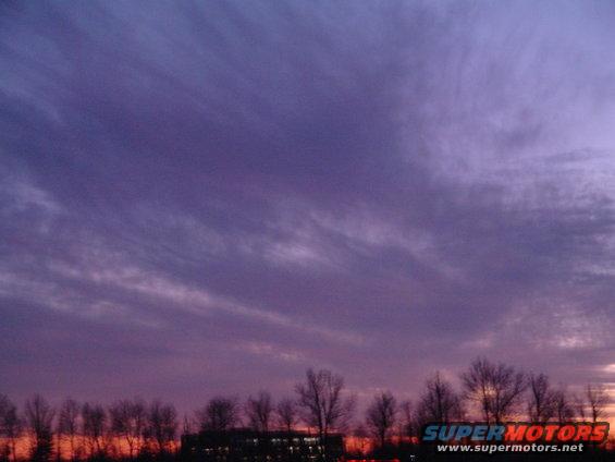 Purple and orange sky at dusk with silhouetted trees and a building below. Supermotors.net logo visible