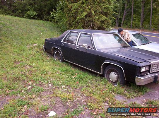 Black sedan parked on grassy shoulder, front passenger side visible. Chrome trim, hubcaps, and grille are present