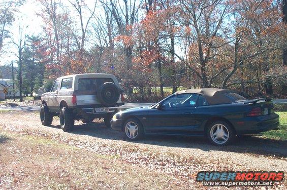 Lifted Ford Bronco towing a dark green convertible on a dirt road