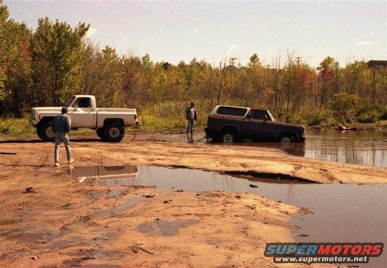 White pickup truck and a dark SUV stuck in a muddy water hole, with two people standing nearby
