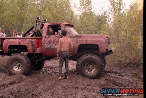 Mud-covered red pickup truck with large tires stuck in mud, people watching
