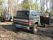 1988 Ford Bronco - A lifted black Ford Bronco with a red and silver stripe is parked in a muddy, wooded area with its driver door open.