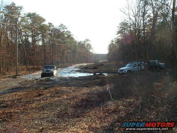 Several off-road vehicles navigating a muddy track through a wooded area with trees and power lines