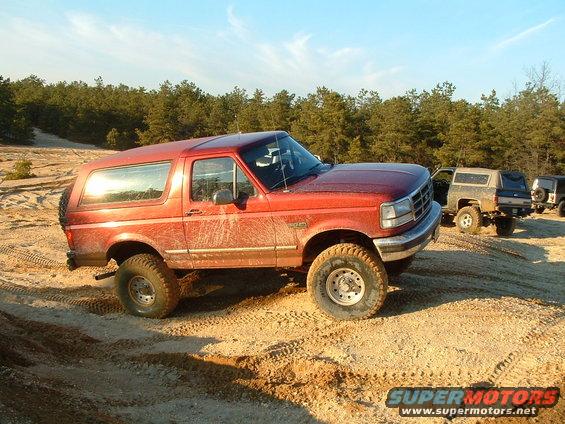 Red Ford Bronco parked on dirt with trees in background. Other SUVs are parked behind it