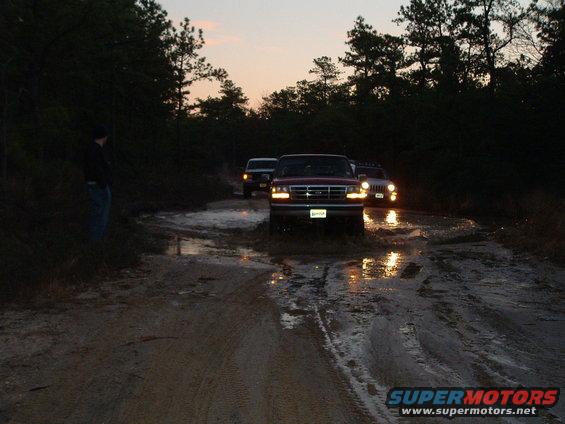 Red pickup truck with grille from 1992 driving through mud puddle at dusk
