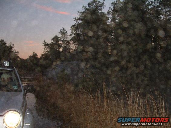 Silver SUV driving on a dirt road at dusk with trees and tall grass