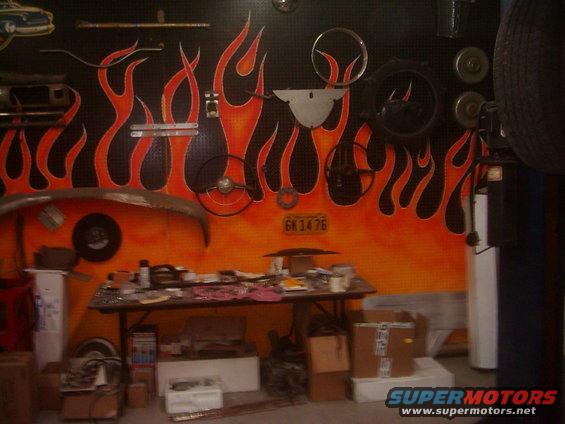 Garage wall with flame mural, steering wheels, car bumper, and a table full of parts. License plate reads GK 1476