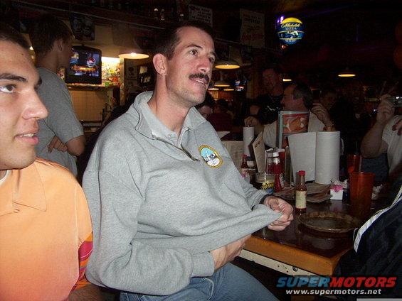 People gathered at a bar, with one man in the foreground pulling his shirt