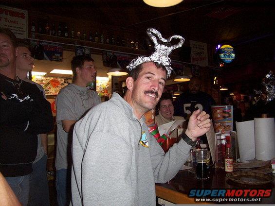 Man with a foil hat on his head smiles at the camera while standing at a bar with other people in the background