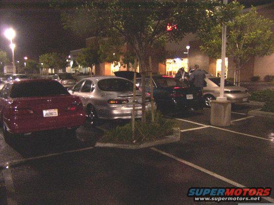 Several cars parked in a lot at night, with people standing near one of them