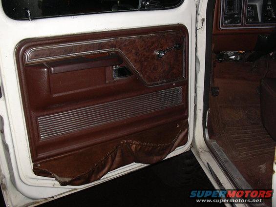 Interior door panel with wood grain trim, chrome handle, and brown upholstery. Dashboard visible on right