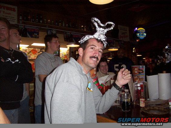 Man with foil hat and mustache smiles at the camera in a bar, with other people in the background