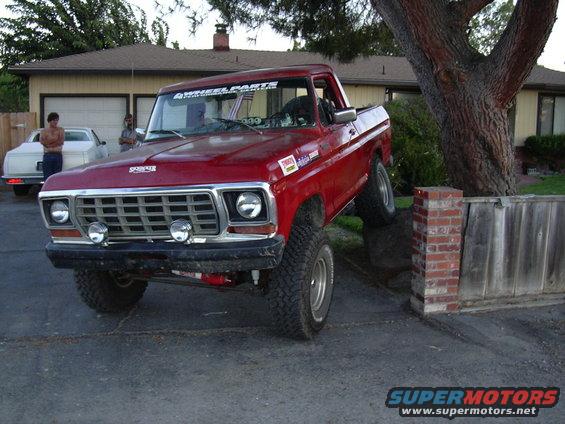 Red pickup truck with lifted suspension, front wheels on a brick wall. White car in background