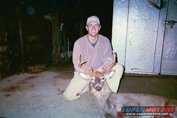 Man kneels with a harvested deer, showing its antlers. A water hose hangs on the wall behind him