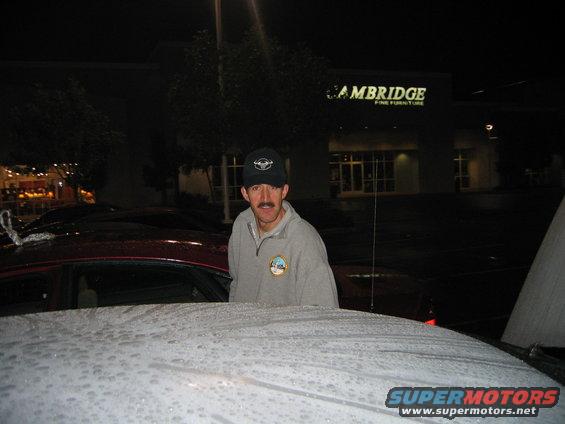 Guy with mustache and baseball cap standing in front of a red car at night. Cambridge Fine Furniture store in background
