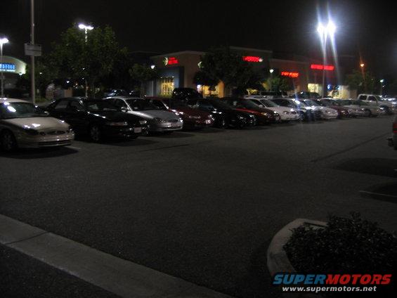 Several cars parked in a lot at night in front of a strip mall
