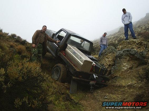 Truck stuck on a steep, rocky incline with three men nearby. A toolbox is in the truck bed