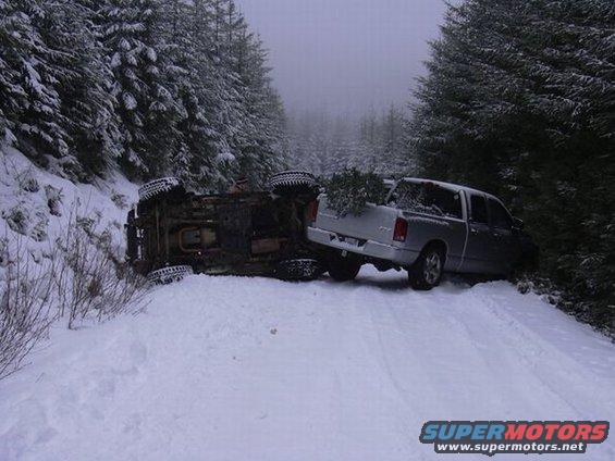 Overturned truck on snowy road next to a silver pickup truck with a Christmas tree in the bed