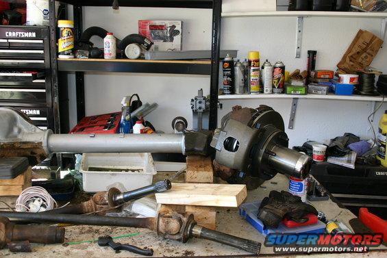 Differential and axle shafts on a workbench with tools and shelves in the background. Supermotors.net logo visible
