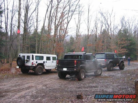 Three SUVs, including a white Hummer and a black pickup, on a muddy dirt road with trees and red flags