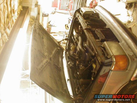 Car engine bay with hood open, showing engine components and hoses. Some tools and garage items are visible in the background