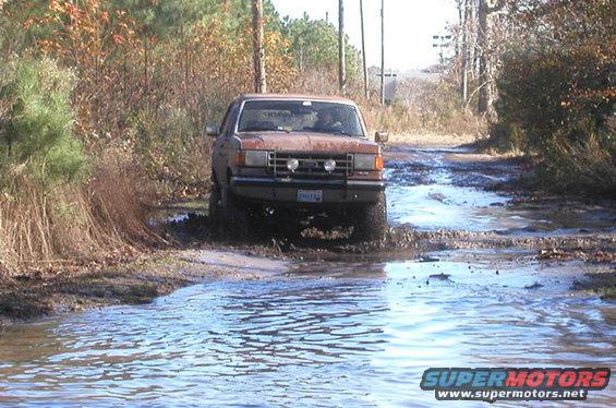 Orange 1987 Ford Bronco driving through a muddy water crossing