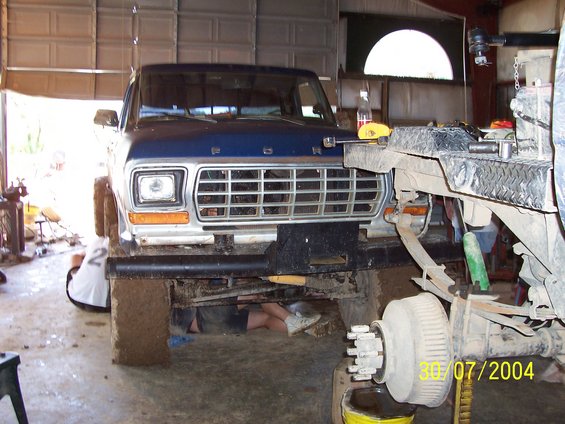 Front view of a blue 1979 Ford F-150 in a garage, with people underneath working on it. Another vehicle is partially visible on the right