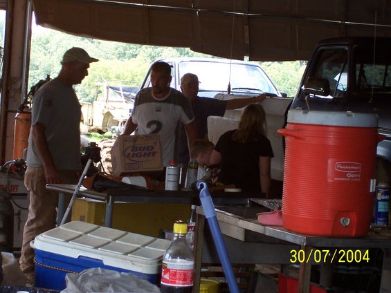 People gathered in a garage or workshop with a cooler, drinks, and two trucks visible in the background