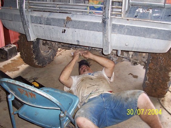 Man lying on concrete floor working under a dirty 4x4 vehicle, blue chair nearby