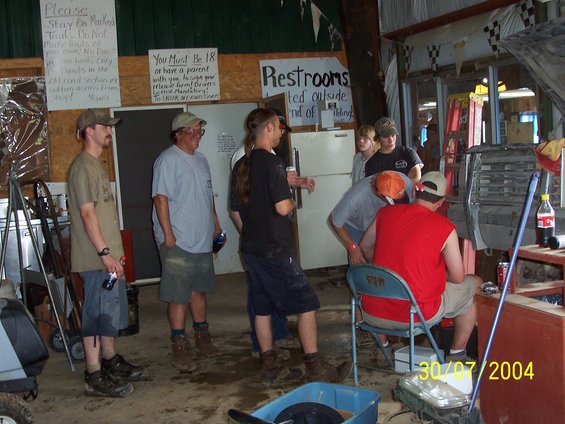 People gathered around a vehicle's front end, possibly working on it. Signs on the wall include 'Please Stay On Marked Trails' and 'Restrooms'