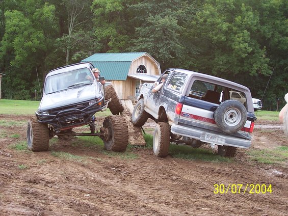 Two modified SUVs, a dark blue Ford Bronco and a silver Toyota, stuck in mud near a barn. Date stamp reads 30/07/2004
