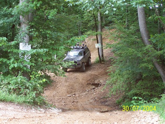Off-road vehicle with roof rack and lights driving up a steep dirt trail in a wooded area