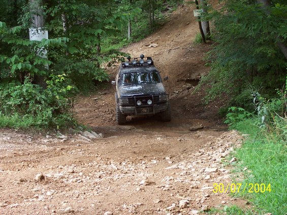 Black SUV with roof lights driving up a steep, rocky dirt path in a wooded area